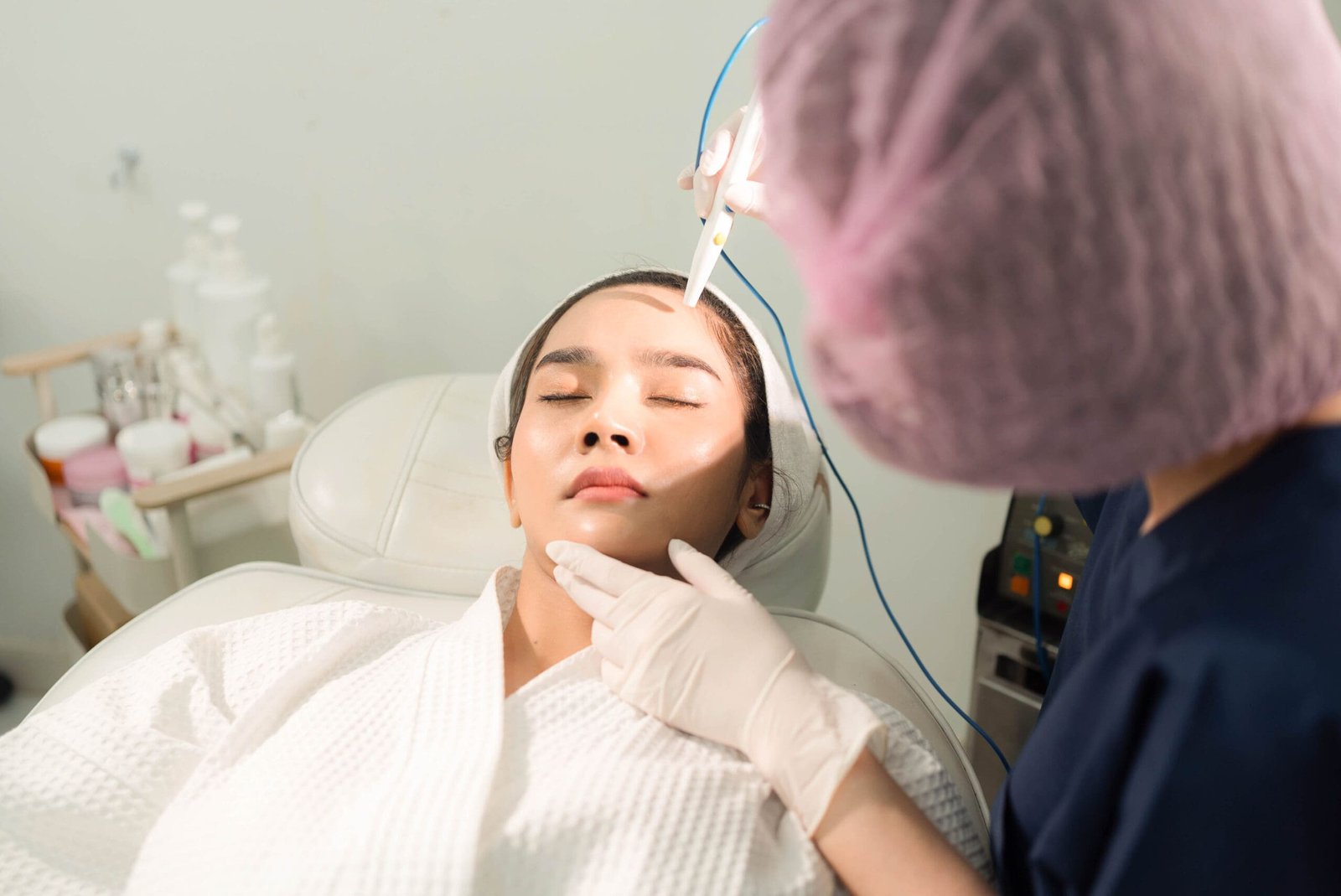 Selective focus of Asian female client in bathrobe and towel on her head lying on bed doing facial peeling procedure with microdermabrasion machine by a blurred beautician in clinic treatment room.