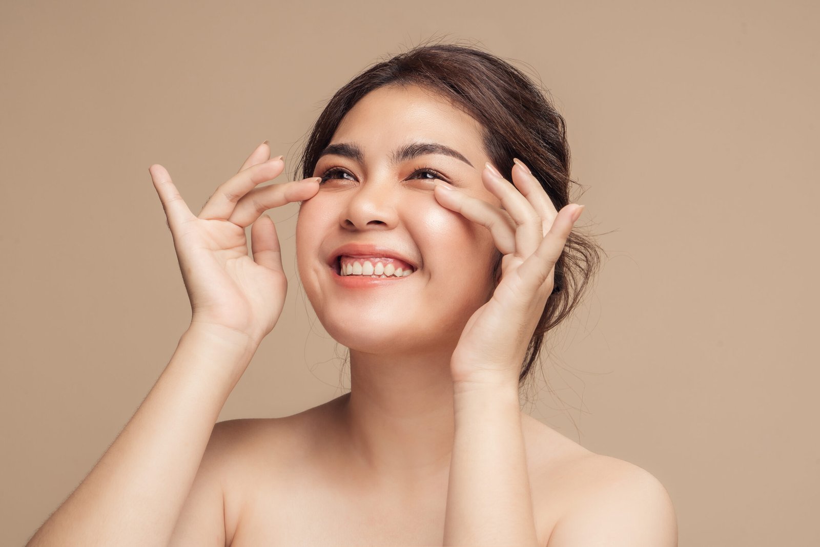 Closeup Asian woman with beauty face touching healthy facial skin portrait. Beautiful smiling Asian female model with natural makeup touching glowing hydrated skin on beige background.